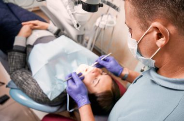 Close up of male dentist in medical mask checking patient teeth. Young woman lying in dental chair while having dental check-up in dental office. Concept of dentistry, stomatology and dental care.