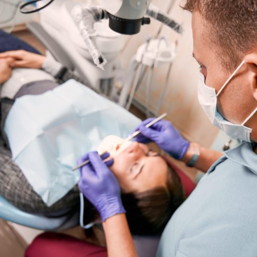 Close up of male dentist in medical mask checking patient teeth. Young woman lying in dental chair while having dental check-up in dental office. Concept of dentistry, stomatology and dental care.