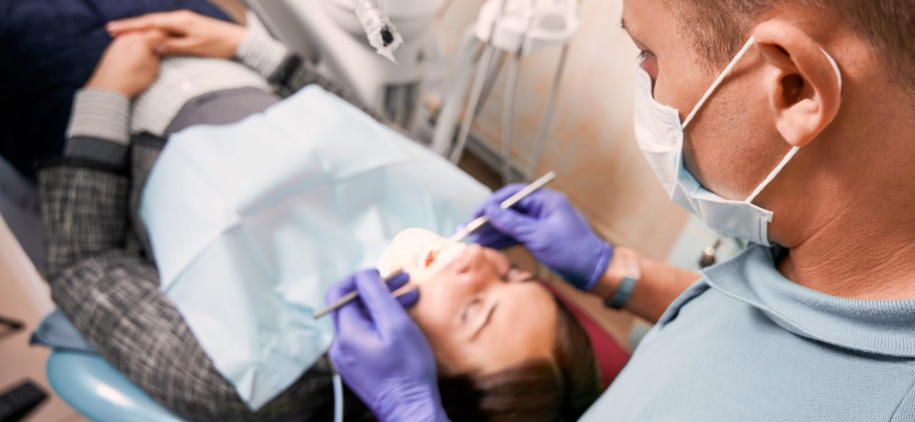Close up of male dentist in medical mask checking patient teeth. Young woman lying in dental chair while having dental check-up in dental office. Concept of dentistry, stomatology and dental care.